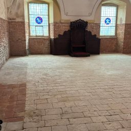 Kamil Rekowski - Sala z ceglanymi ścianami, kamienną podłogą i ozdobnym krzesłem. Widoczne okna z witrażami i fragment sufitu z żyrandolem. Wnętrze w trakcie prac wykończeniowych.