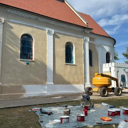Kamil Rekowski - Malowanie elewacji kościoła w trakcie realizacji. Widoczny podnośnik koszowy, farby i narzędzia malarskie rozłożone na trawie. Częściowo pomalowana ściana budynku.
