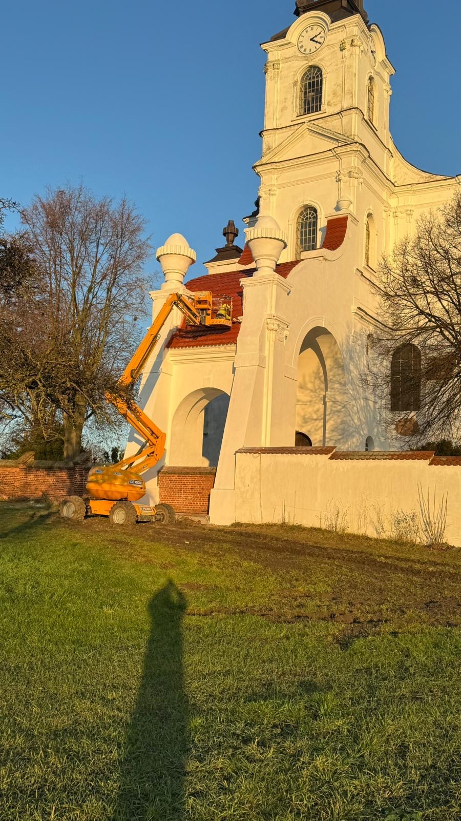 Prace na dachu kościoła z użyciem podnośnika koszowego. Widoczna biała fasada budynku i czerwona dachówka. W tle drzewa i błękitne niebo.