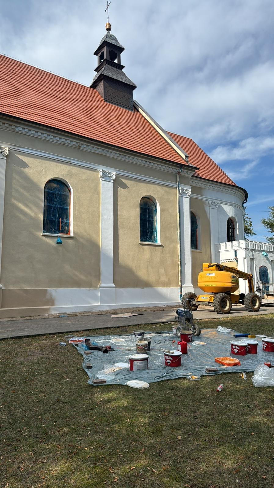 Malowanie elewacji kościoła w trakcie realizacji. Widoczny podnośnik koszowy, farby i narzędzia malarskie rozłożone na trawie. Częściowo pomalowana ściana budynku.