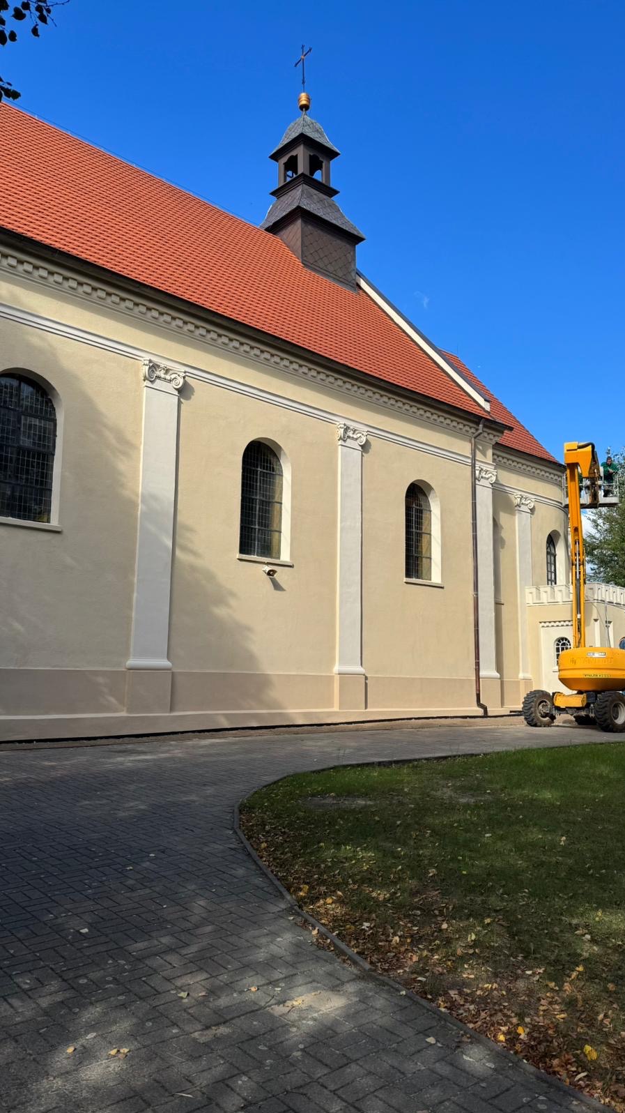 Odnowiona elewacja kościoła w beżowej tonacji, z widoczną podnośnikiem koszowym i fragmentem chodnika z kostki brukowej. Czerwony dach i błękitne niebo w tle.