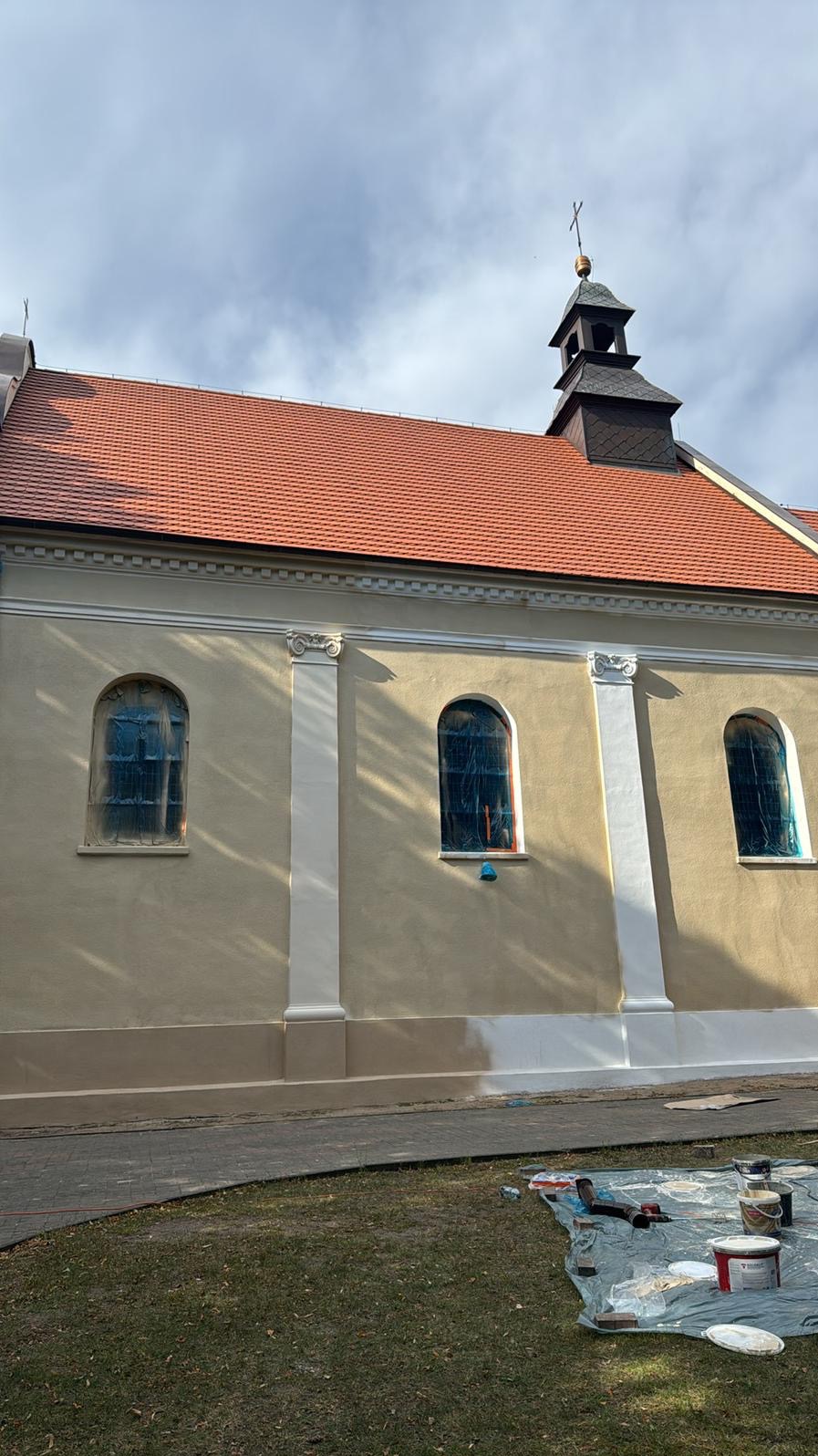 Elewacja budynku sakralnego w trakcie odnawiania, z widocznymi zabezpieczonymi oknami i częściowo pomalowaną dolną partią ściany. Na pierwszym planie rozłożona plandeka z farbami.