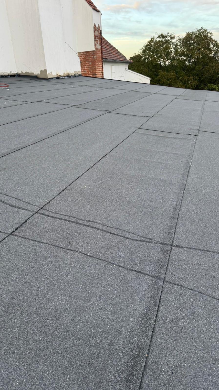 Szary dach pokryty papą termozgrzewalną w kwadraty, widok z góry na budynek z ceglanym kominem i zielenią w tle, pochmurne niebo.