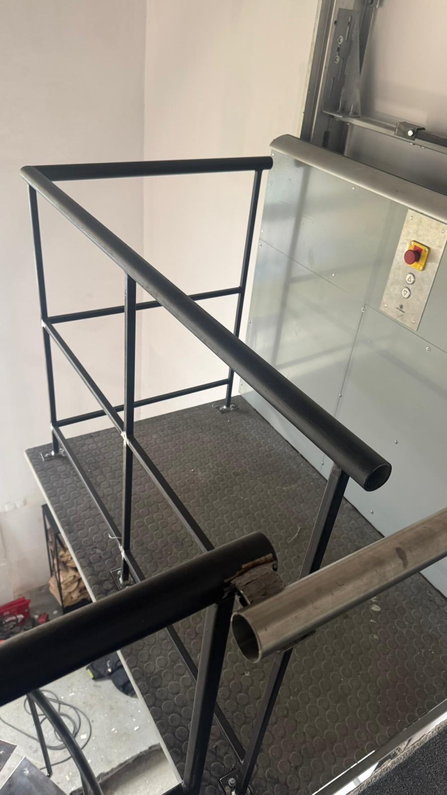 Metalowa konstrukcja schodów z czarną poręczą i szarą platformą, widok z góry, częściowo widoczny mechanizm windy.