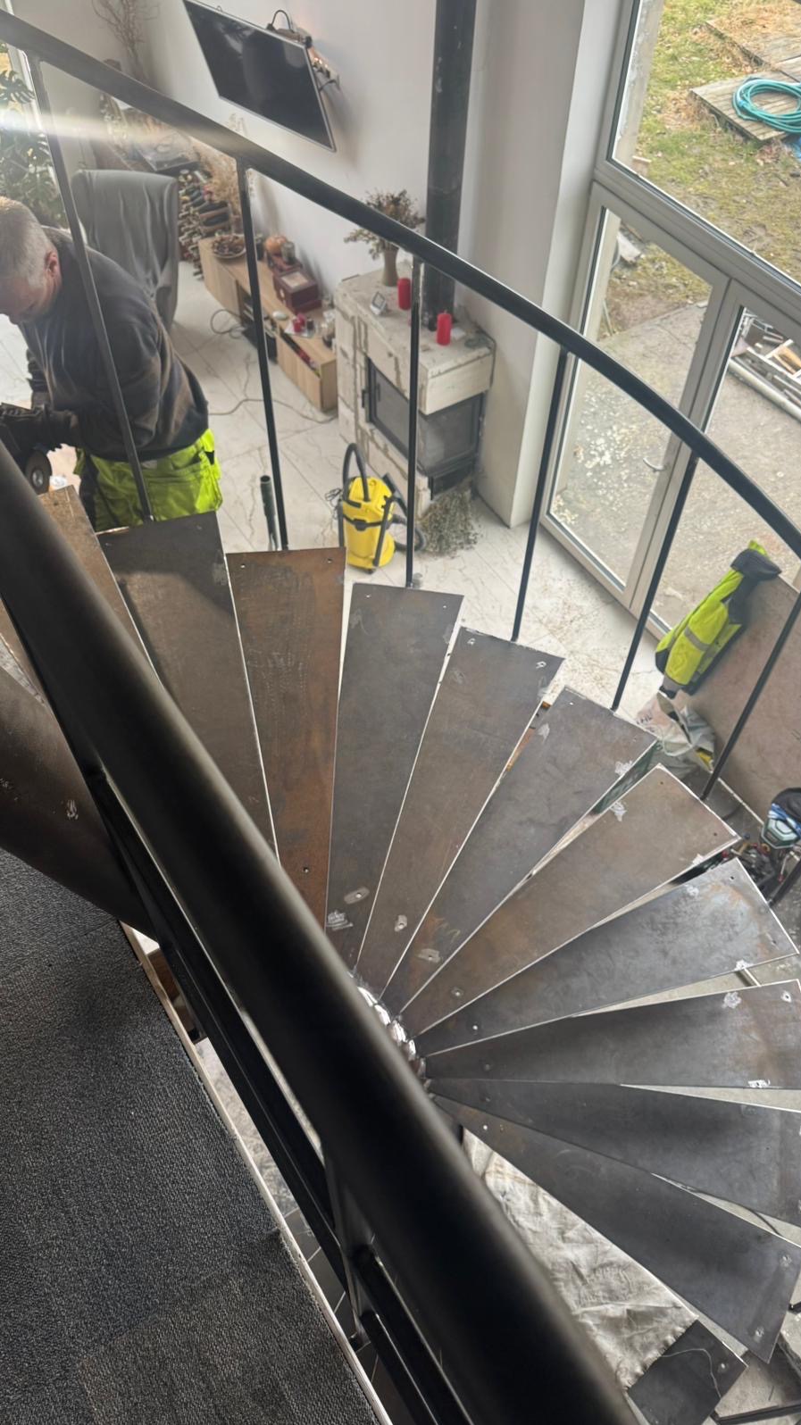 Montaż metalowych schodów spiralnych w Olzie. Widok z góry na surowe stopnie i pracownika w odblaskowej kamizelce podczas szlifowania.