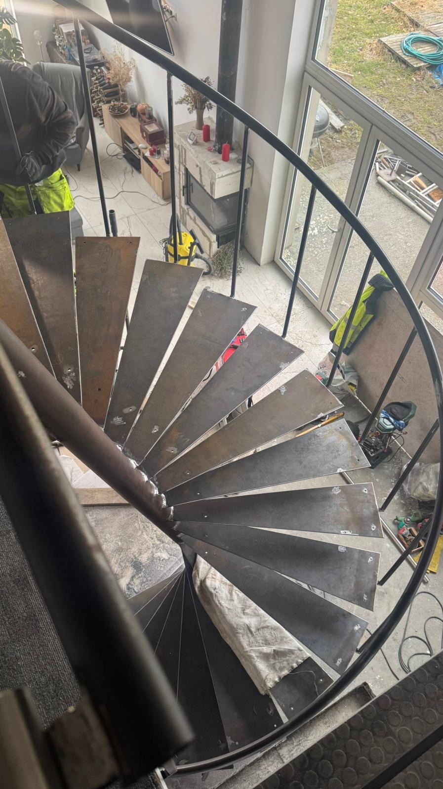 Metalowe schody spiralne w trakcie montażu, widok z góry. Surowe stopnie, czarna balustrada, w tle okno i wyposażenie wnętrza. Widoczne ślady prac montażowych.