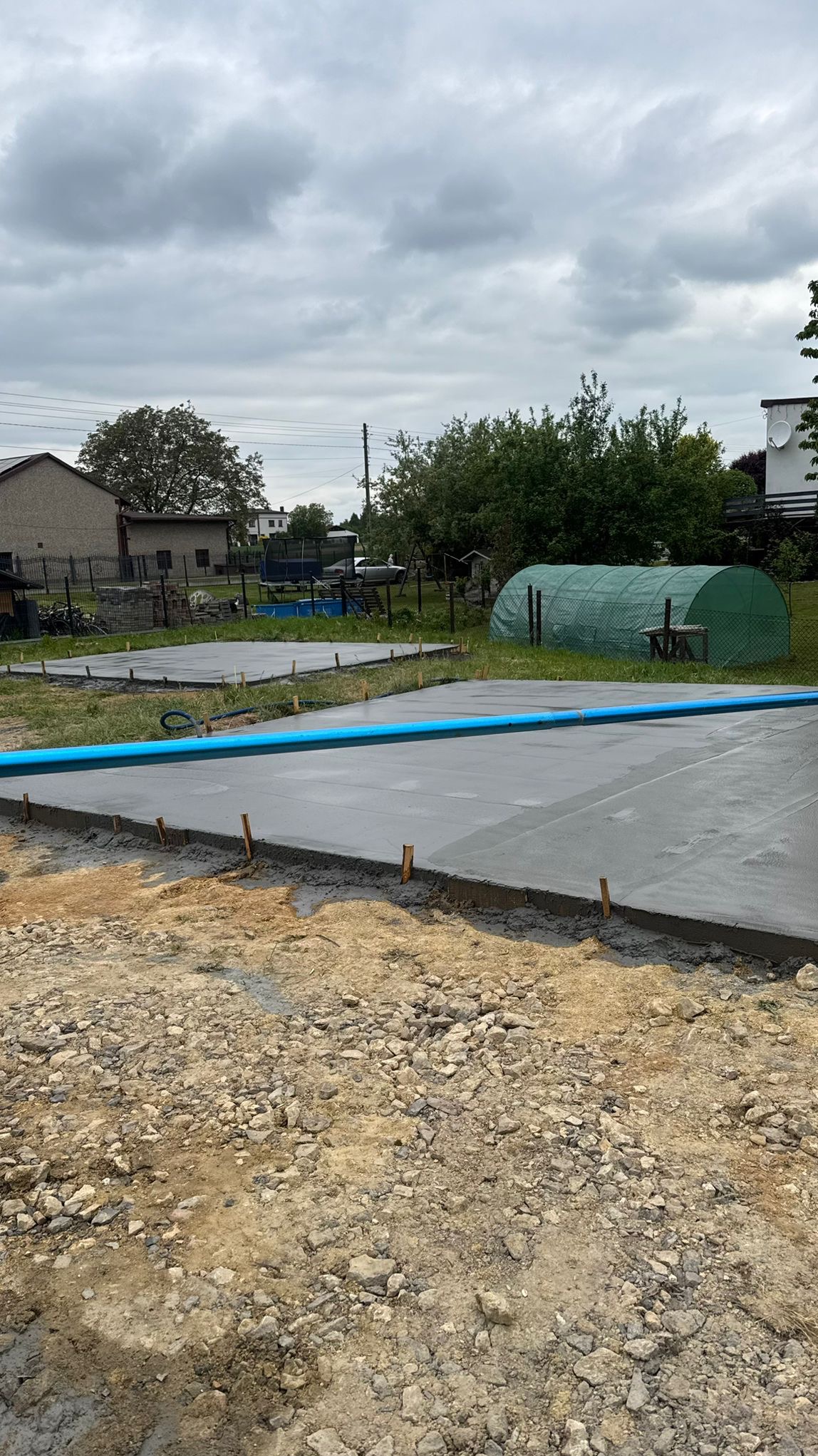 Wylana, świeża betonowa podłoga na zewnątrz, z widocznymi drewnianymi kołkami i niebieską rurą. W tle budynek, drzewa i szklarnia pod pochmurnym niebem.