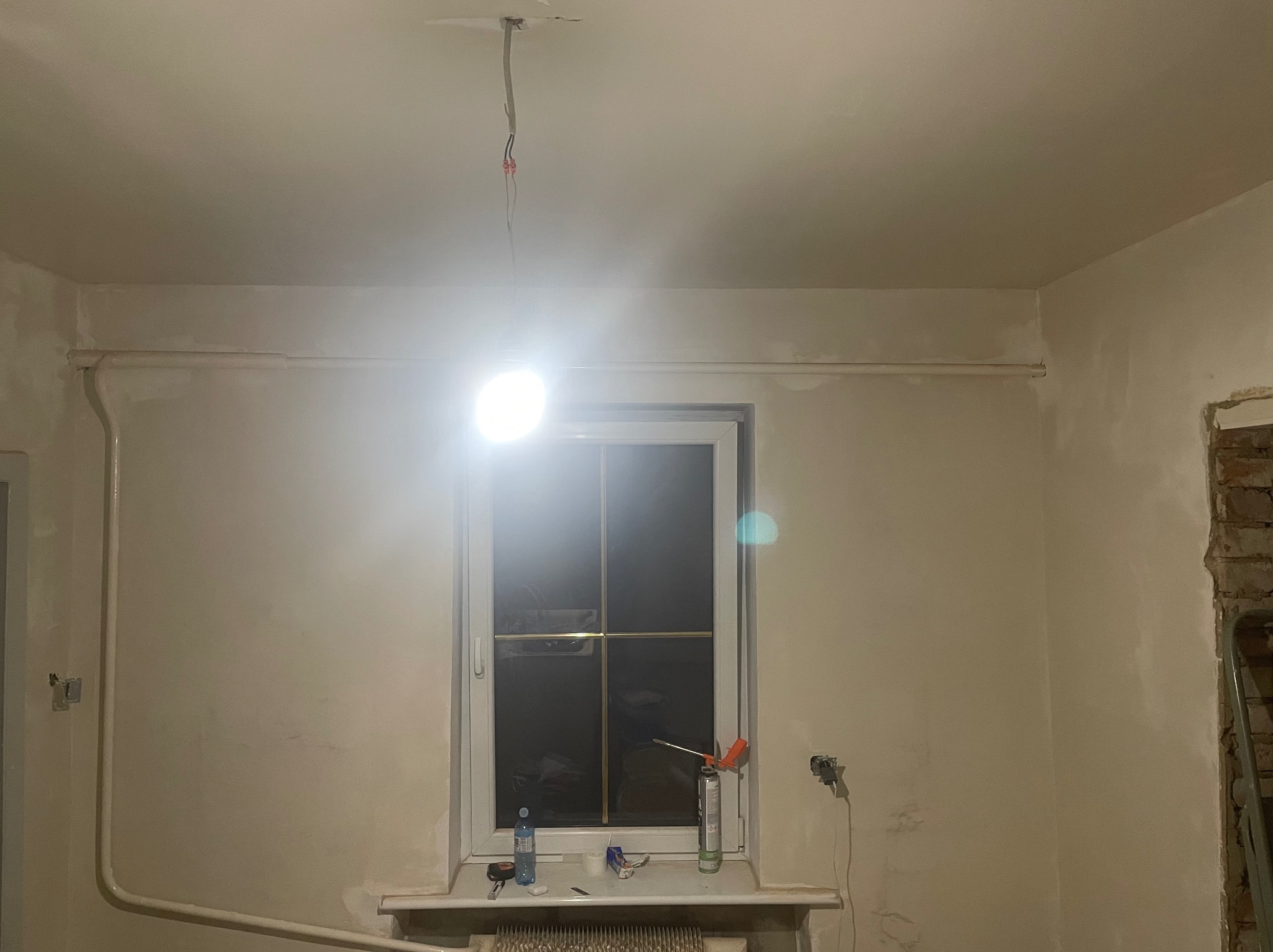 Ściana po szpachlowaniu w Kaliszu, widoczne rury, okno z narzędziami i wystające kable z sufitu. Ściana przygotowana do dalszych prac wykończeniowych.