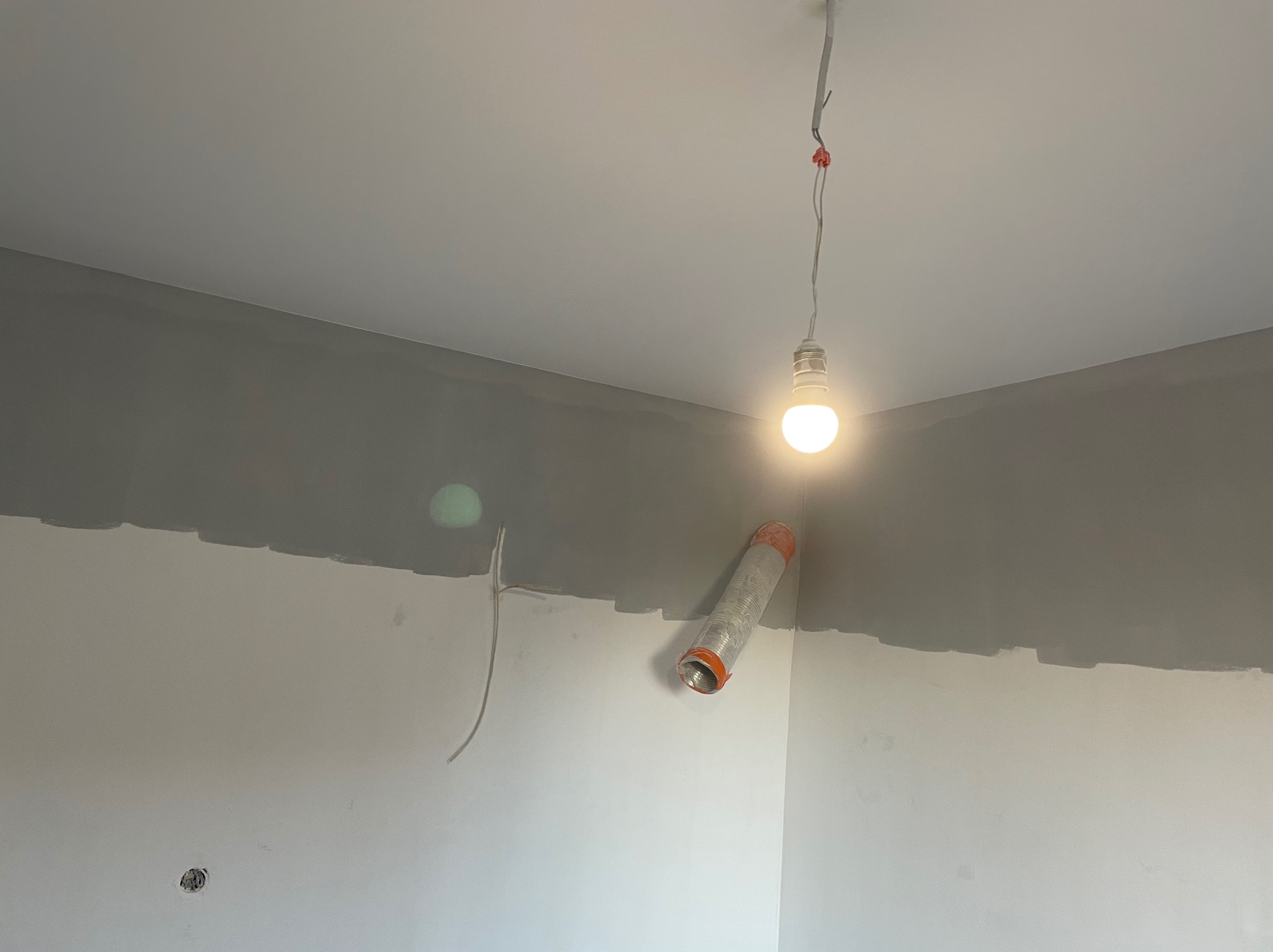 Ściana w trakcie malowania na szaro, widoczne przewody elektryczne i tymczasowe oświetlenie żarówką. Częściowo pomalowana ściana, widoczny przewód wentylacyjny.