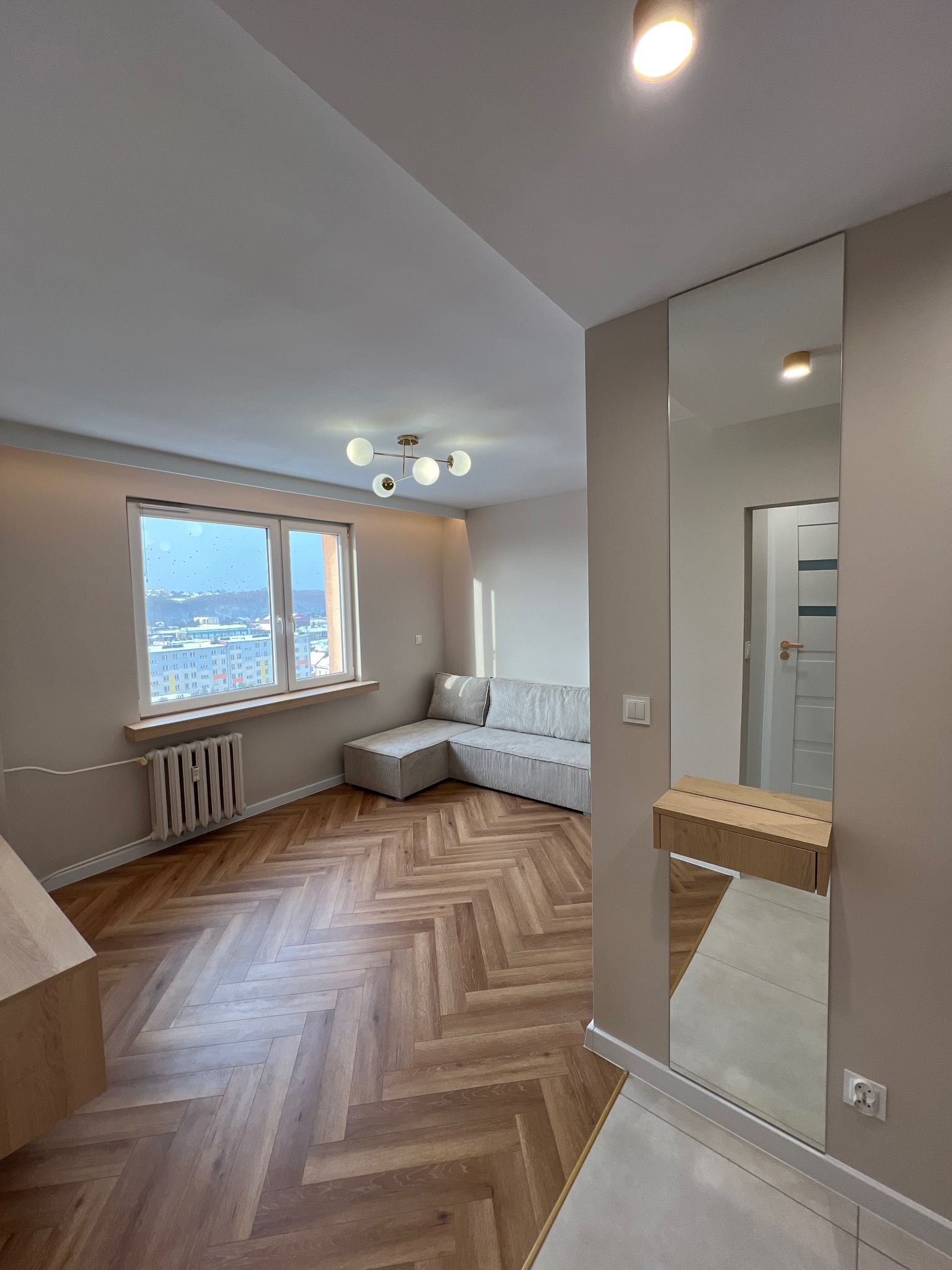 Jasne wnętrze po remoncie: parkiet w jodełkę, beżowe ściany, okno z widokiem na miasto. Minimalistyczny design z lustrem i drewnianą konsolą.