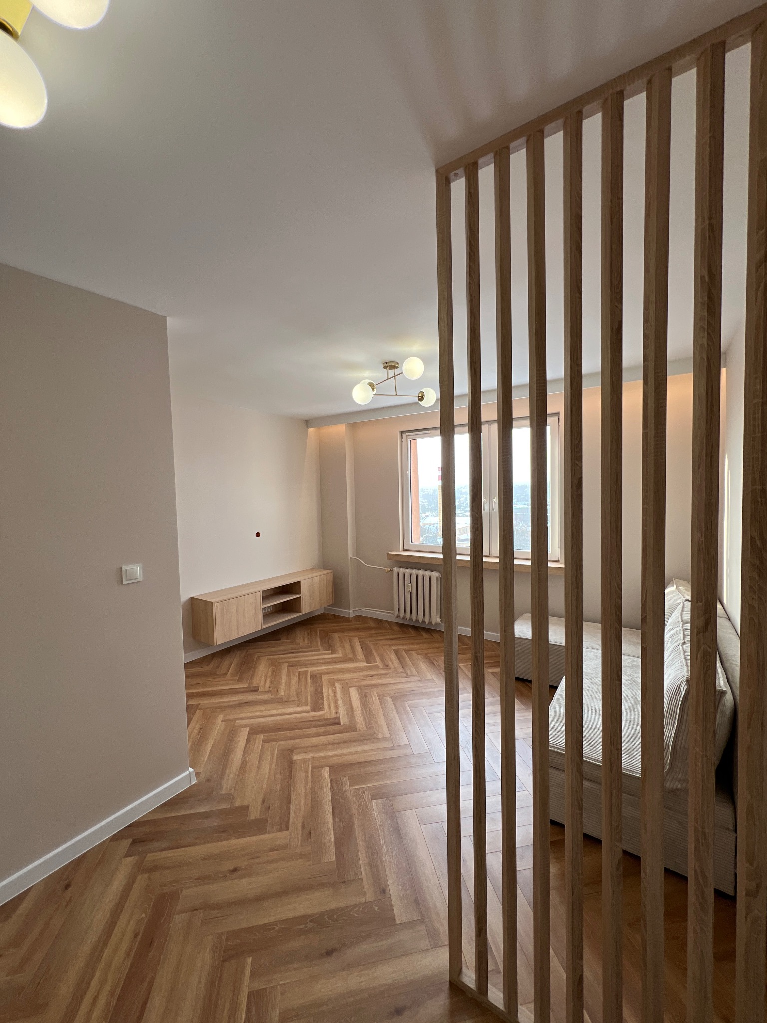 Jasne wnętrze po remoncie: parkiet w jodełkę, beżowe ściany, drewniana ażurowa ścianka działowa. Minimalistyczna szafka RTV i nowoczesna lampa sufitowa.