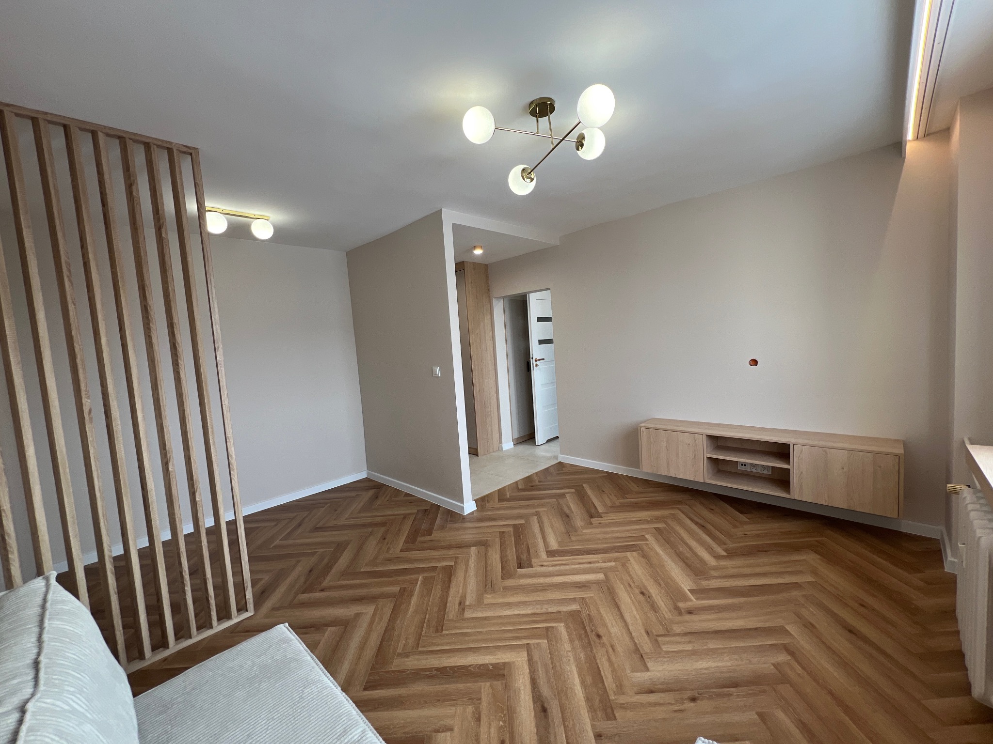 Jasne wnętrze po remoncie: parkiet w jodełkę, drewniana zabudowa TV, dekoracyjna ścianka działowa z listewek i nowoczesna lampa sufitowa.