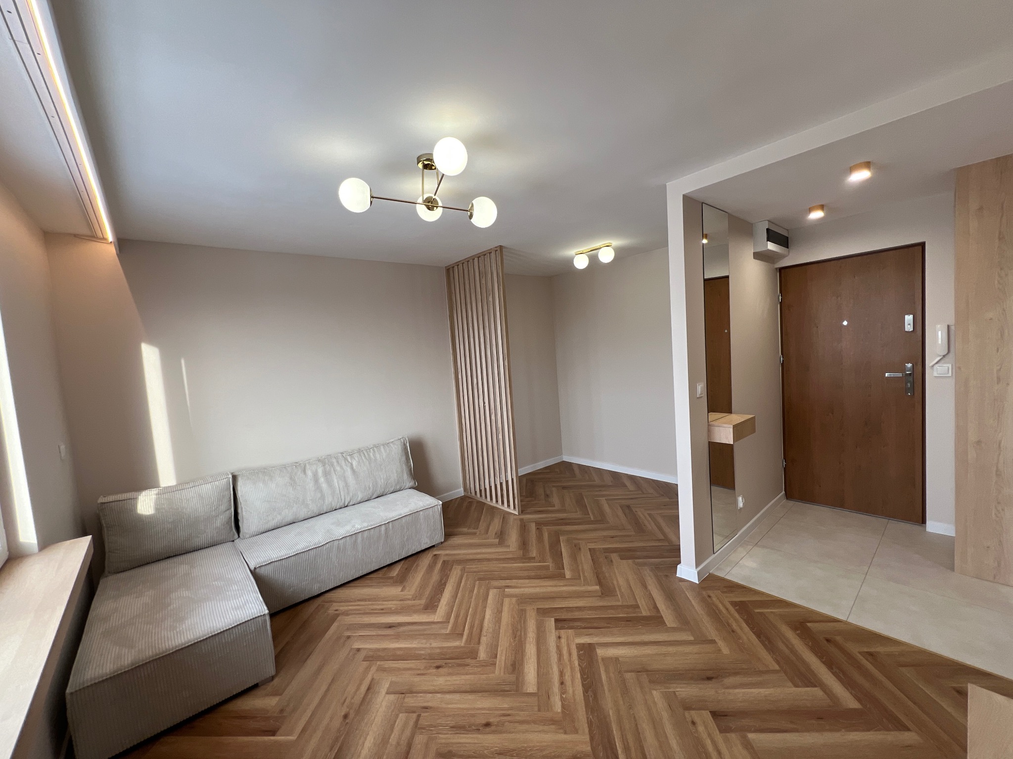 Jasne wnętrze po remoncie: parkiet w jodełkę, beżowe ściany, sofa z tkaniny w prążki, ażurowa ścianka działowa i nowoczesne oświetlenie.