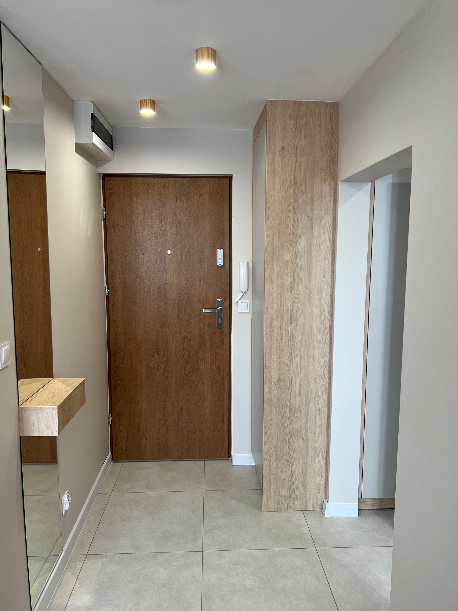 Nowoczesny przedpokój z drewnianymi akcentami: drzwi, szafa i konsola pod lustrem. Beżowe ściany i płytki podłogowe. Dwa okrągłe plafony oświetlają przestrzeń.