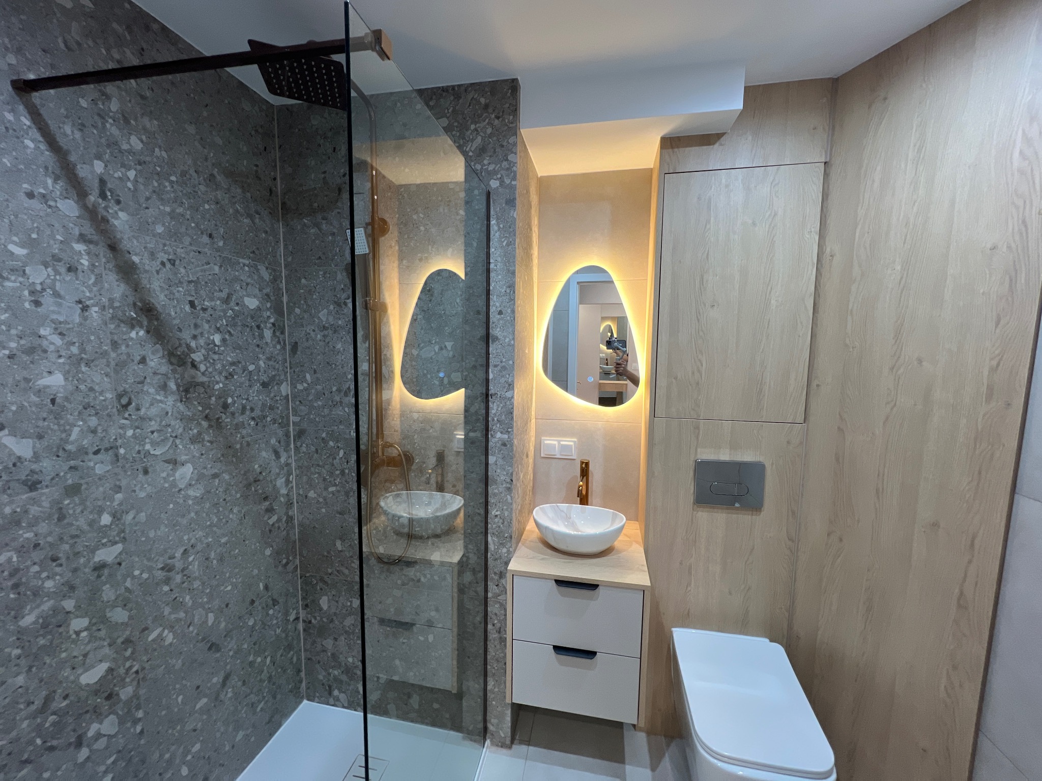 Nowoczesna łazienka z kabiną prysznicową, umywalką na szafce i podświetlanym lustrem. Ściany wykończone płytkami imitującymi kamień, drewnopodobne panele na ścianie.