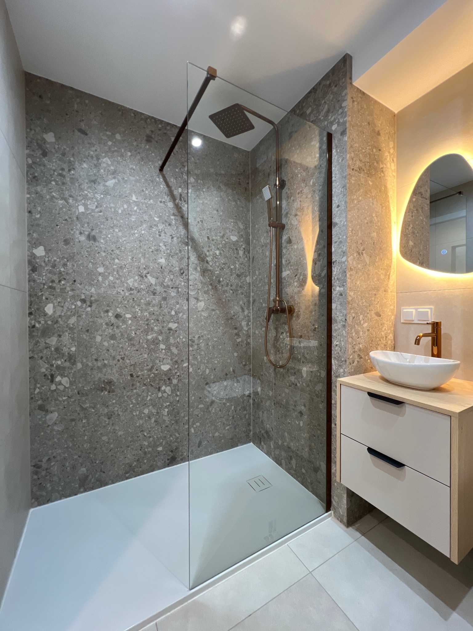Nowoczesna łazienka z kabiną prysznicową typu walk-in, szarą mozaiką na ścianach i umywalką nablatową z podświetlanym lustrem. Armatura w kolorze miedzi.