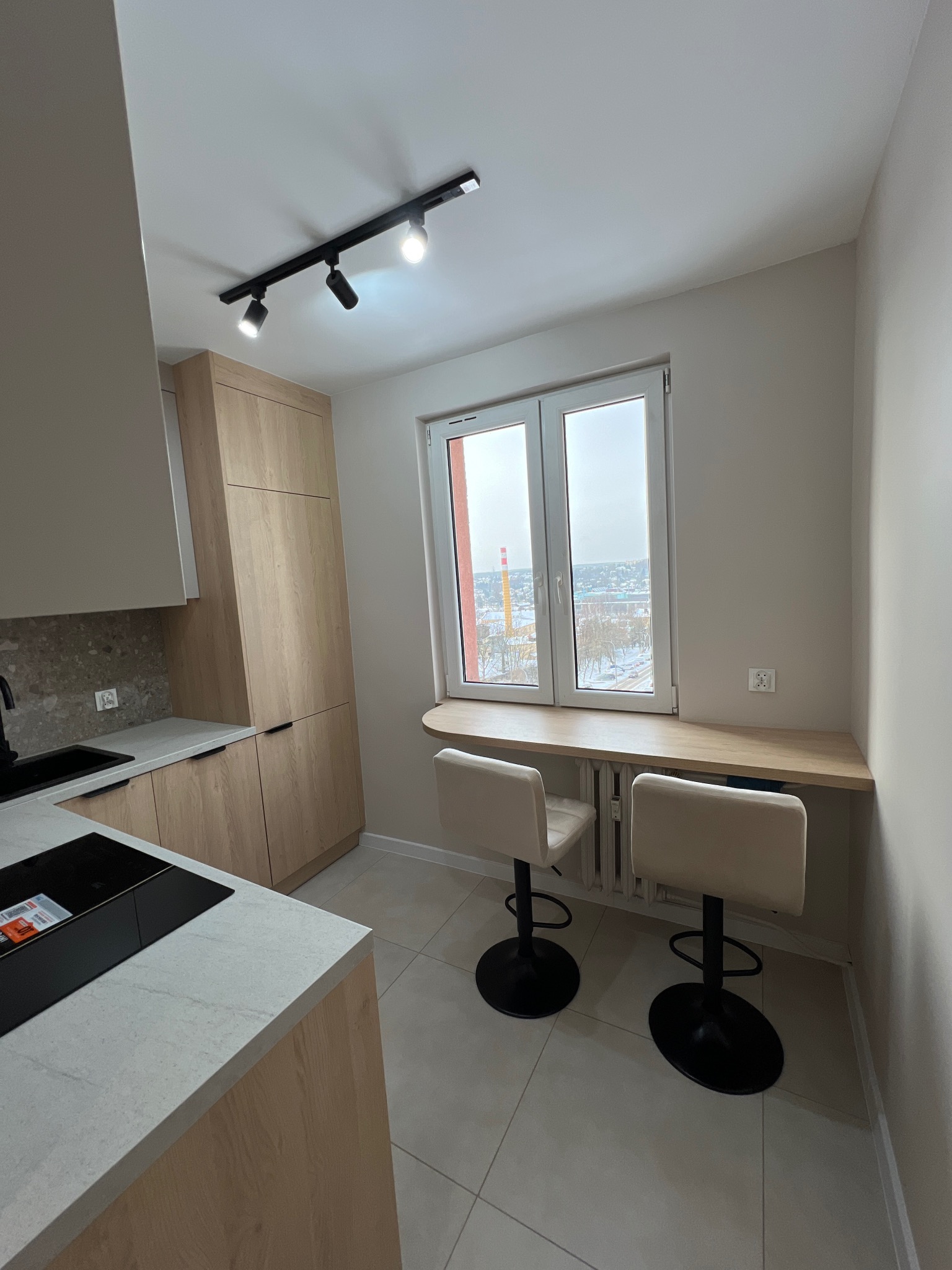 Jasna kuchnia z drewnianymi szafkami, blatem z kamienia i oknem z barkiem. Dwa krzesła barowe stoją przy oknie. Minimalistyczne oświetlenie sufitowe.