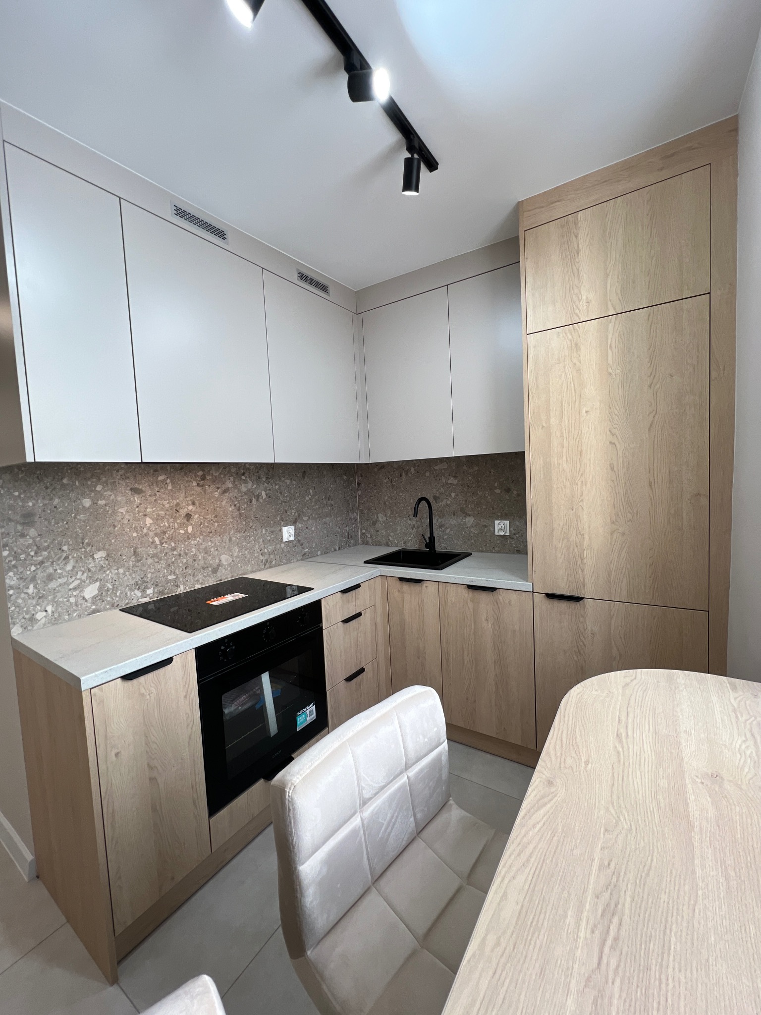Nowoczesna kuchnia z drewnianymi i białymi szafkami, czarną armaturą i blatem z kamienia. Oświetlenie punktowe podkreśla minimalistyczny design.