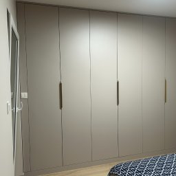 Kastero - Szafa wnękowa z frontami w kolorze taupe, z długimi, smukłymi uchwytami w odcieniu złota, wkomponowana w jasne wnętrze sypialni, widoczny fragment łóżka.