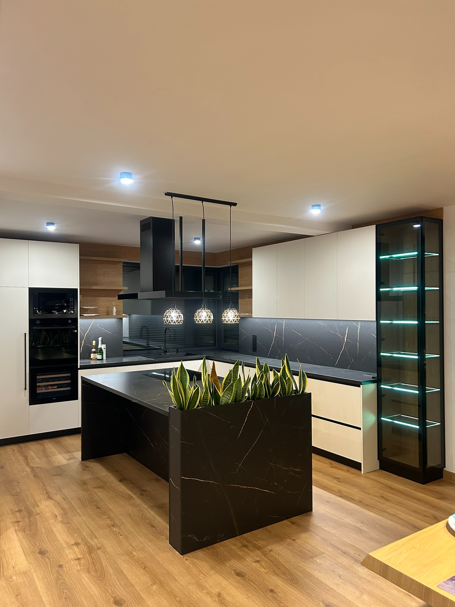 Nowoczesna kuchnia z wyspą i marmurowym blatem w Ostrowie. Białe szafki górne kontrastują z czarnymi elementami. Oświetlenie punktowe i przeszklona witryna.