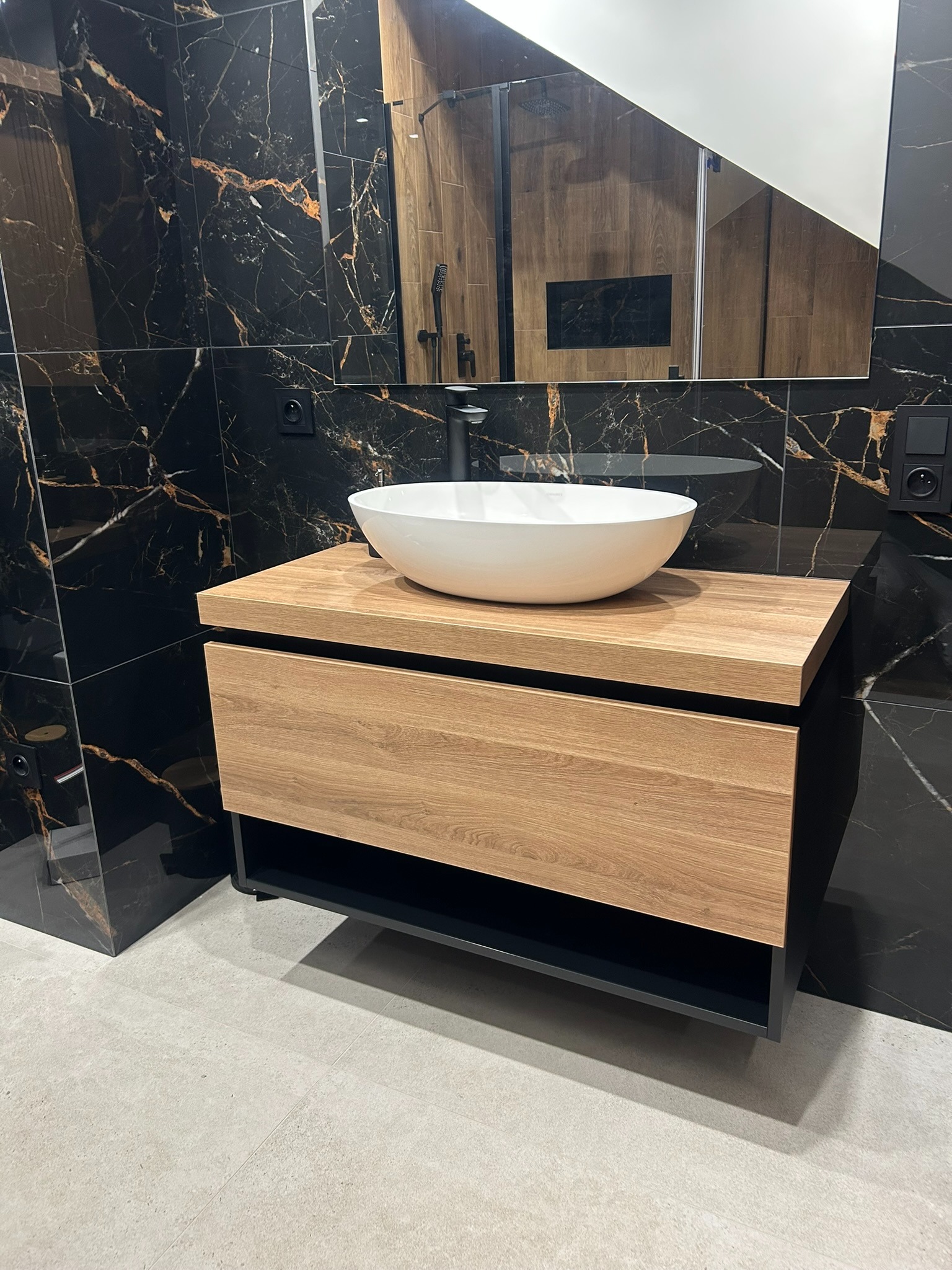 Nowoczesna szafka łazienkowa z drewnianym blatem i białą umywalką na tle czarnych płytek imitujących marmur. Widoczna armatura i fragment kabiny prysznicowej.