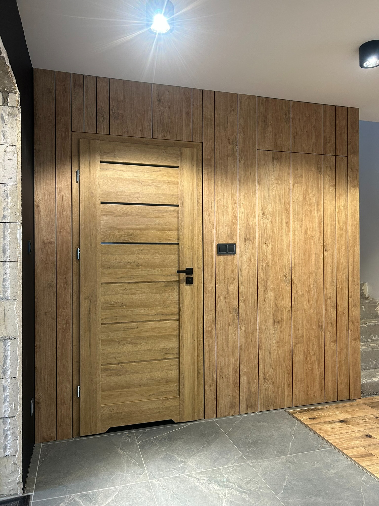 Drewniana zabudowa ściany z ukrytymi drzwiami i szafką, wykończona w pionowe panele. Szare płytki podłogowe i oświetlenie sufitowe.