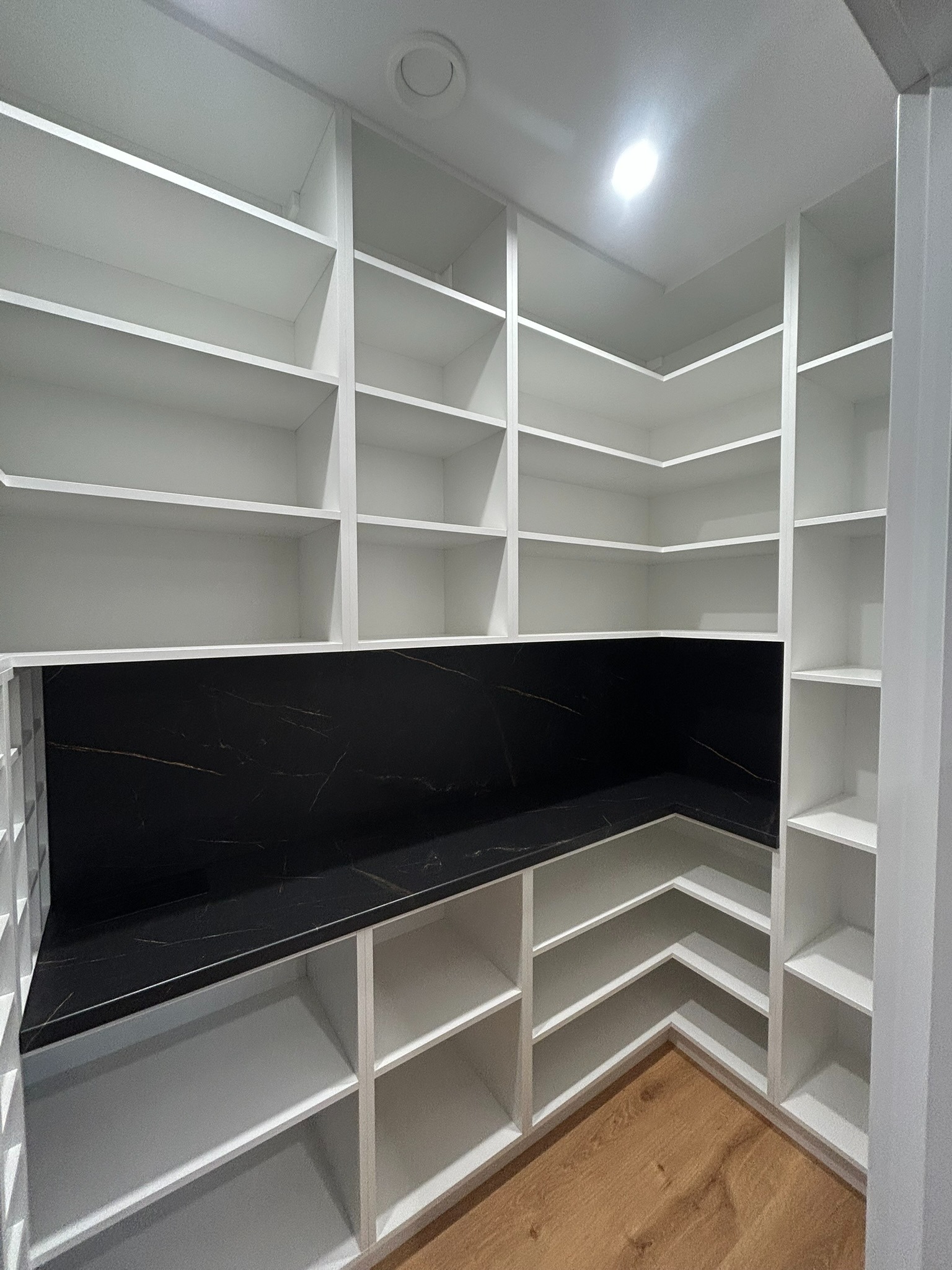 Biała, narożna garderoba z półkami i czarnym blatem z efektem marmuru, oświetlona od góry. Widoczna drewniana podłoga.