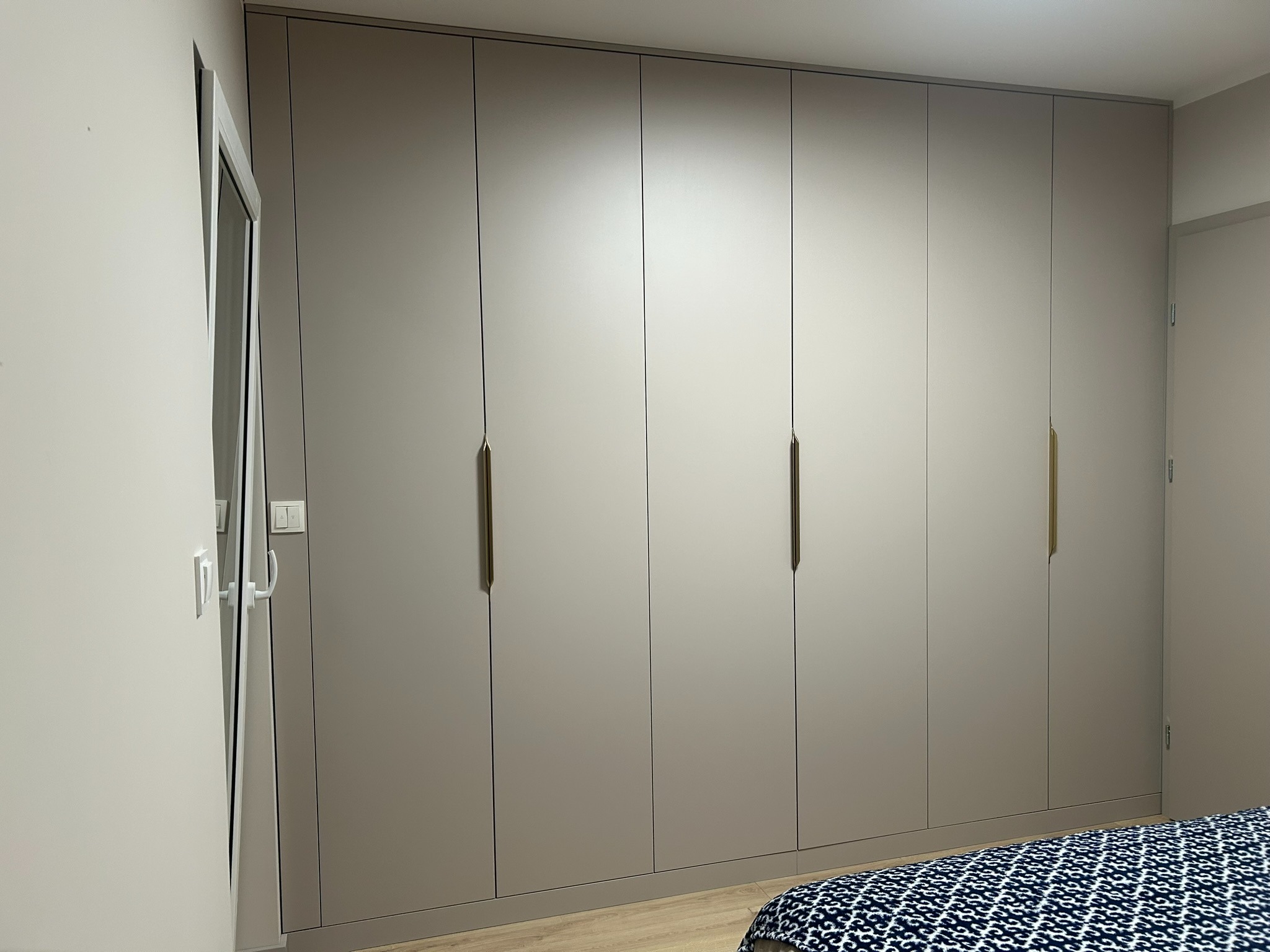Szafa wnękowa z frontami w kolorze taupe, z długimi, smukłymi uchwytami w odcieniu złota, wkomponowana w jasne wnętrze sypialni, widoczny fragment łóżka.