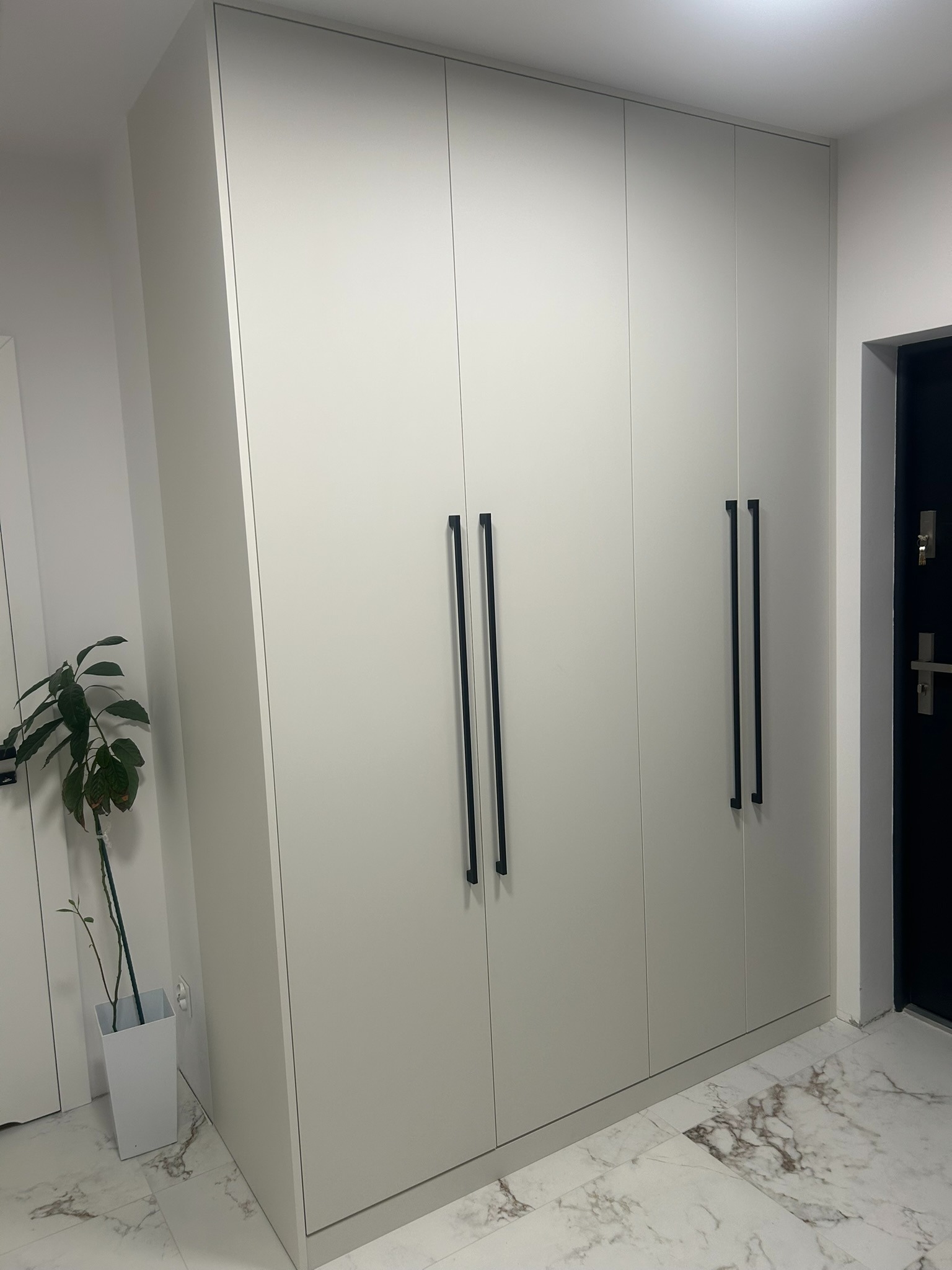Jasnoszara szafa na wymiar z długimi, czarnymi uchwytami, stojąca w przedpokoju z marmurową podłogą i rośliną w białej donicy. Minimalistyczny design.