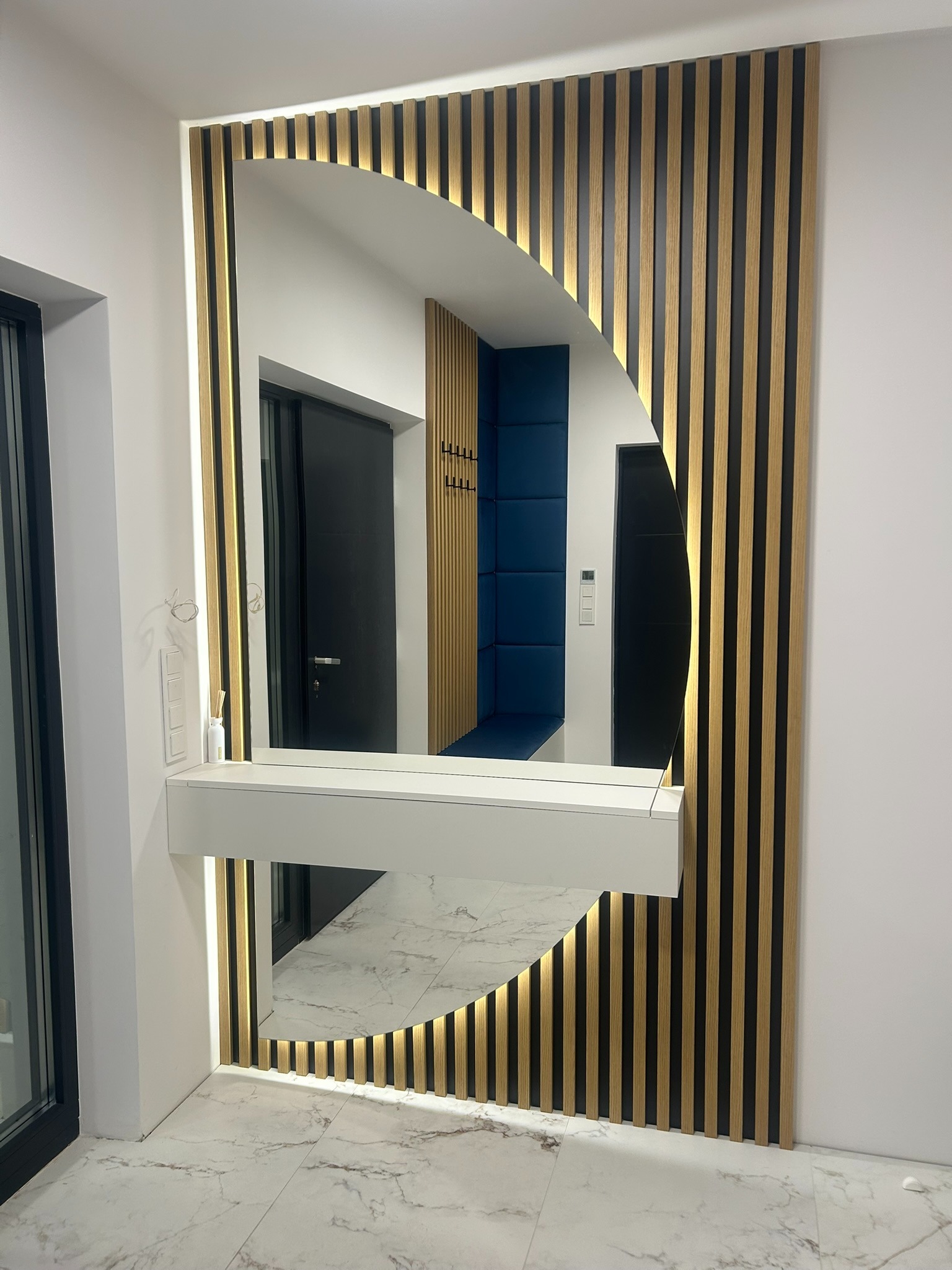 Nowoczesna aranżacja przedpokoju z lustrem w kształcie półkola, obudowanym drewnianymi lamelami z podświetleniem LED i białą konsolą. Widoczne drzwi i marmurowa podłoga.