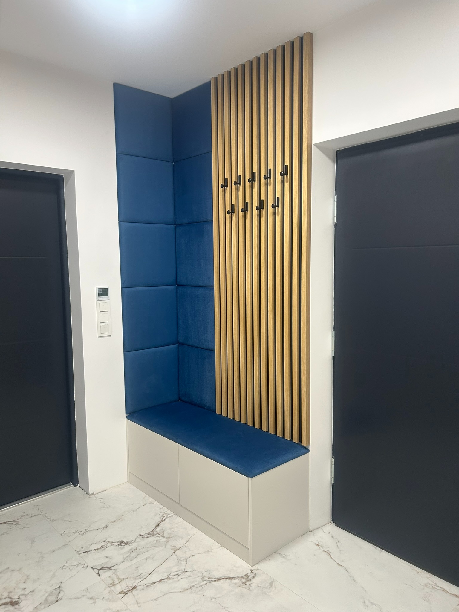 Narożna zabudowa w przedpokoju: tapicerowany panel w odcieniach niebieskiego, drewniane lamele z wieszakami i szafka z siedziskiem. Nowoczesny design.