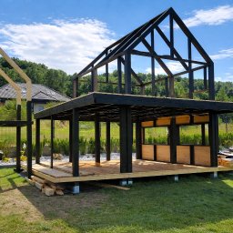OGRODOMY SPÓŁKA Z OGRANICZONĄ ODPOWIEDZIALNOŚCIĄ - Wznoszona konstrukcja drewniana z czarnymi elementami, prawdopodobnie pergola lub domek letniskowy, na tle zieleni i błękitnego nieba. Widoczne detale konstrukcyjne i etap budowy.