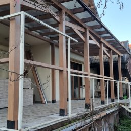 OGRODOMY SPÓŁKA Z OGRANICZONĄ ODPOWIEDZIALNOŚCIĄ - Zadaszony taras z drewnianą konstrukcją i białymi barierkami, widok z dołu. Elementy konstrukcyjne w kolorze brązowym. W tle budynek z oknami i roletami.