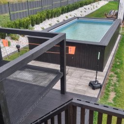 OGRODOMY SPÓŁKA Z OGRANICZONĄ ODPOWIEDZIALNOŚCIĄ - Czarny, drewniany taras z siatką i schodami prowadzący do basenu w ogrodzie. Widok z góry na konstrukcję i otoczenie z zielenią.