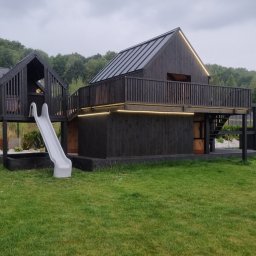 OGRODOMY SPÓŁKA Z OGRANICZONĄ ODPOWIEDZIALNOŚCIĄ - Nowoczesny, dwupoziomowy plac zabaw z drewna w kolorze antracytu, z oświetleniem LED, zjeżdżalnią i piaskownicą, na tle zielonego trawnika i drzew.