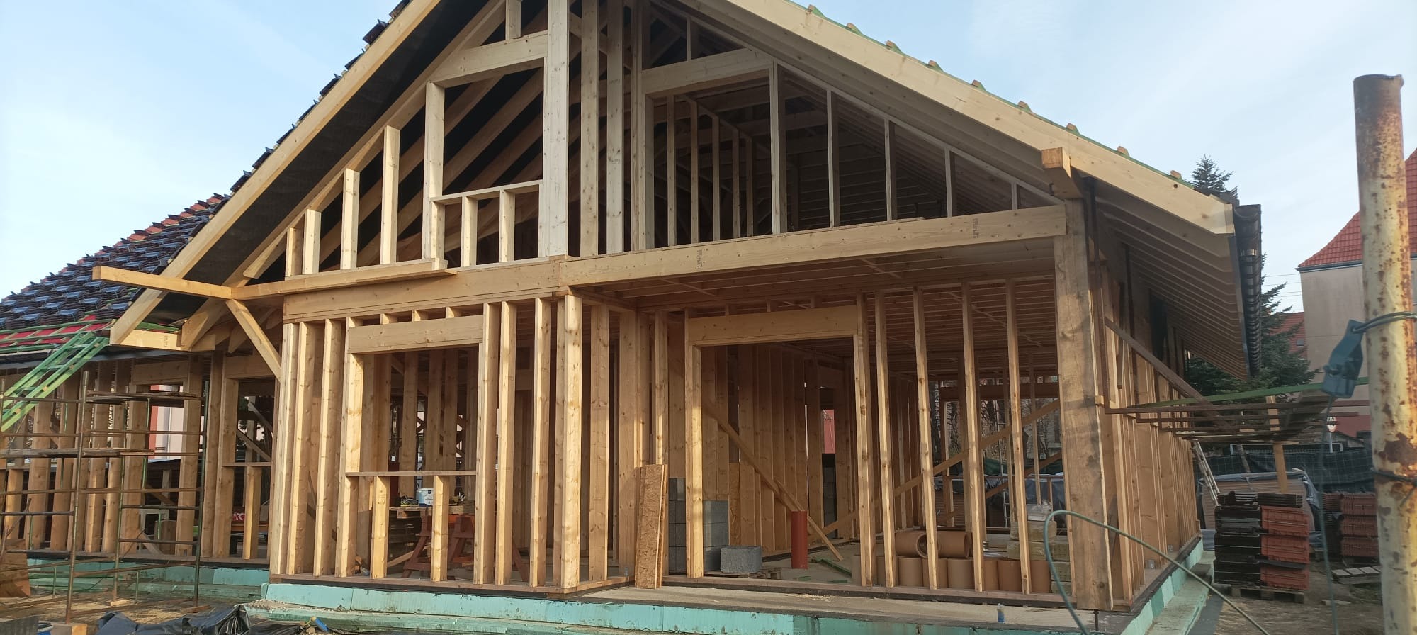 Konstrukcja domu w technologii szkieletowej w trakcie budowy. Widoczne drewniane ramy ścian, okien i dachu. Częściowo położone dachówki.