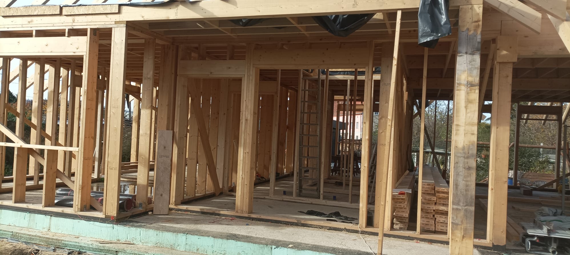 Konstrukcja domu szkieletowego w trakcie budowy, widoczne drewniane ramy ścian i stropu, z zewnątrz widok na drzewa i budynki.