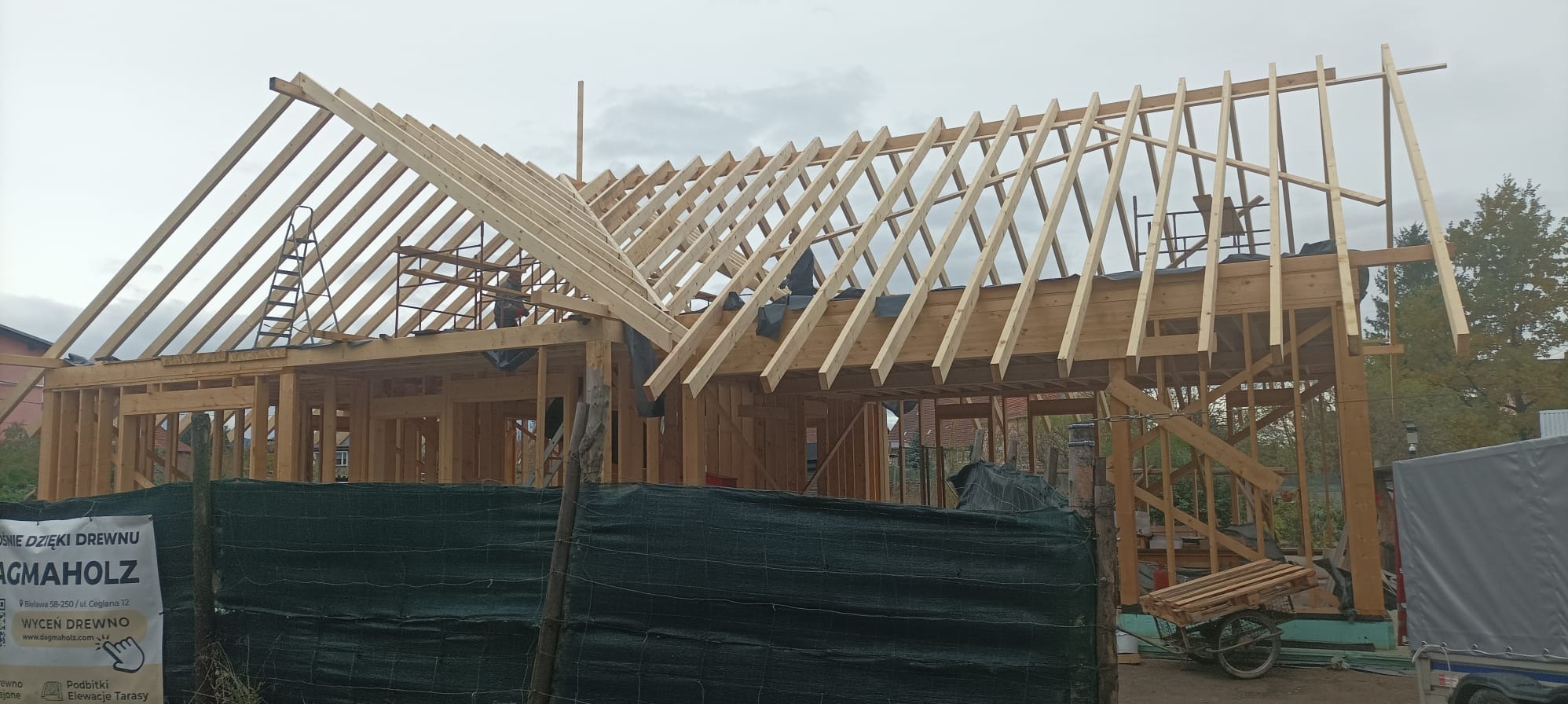 Konstrukcja domu szkieletowego w trakcie budowy, widoczne drewniane elementy nośne i więźba dachowa. Na pierwszym planie siatka ogrodzeniowa i reklama firmy.