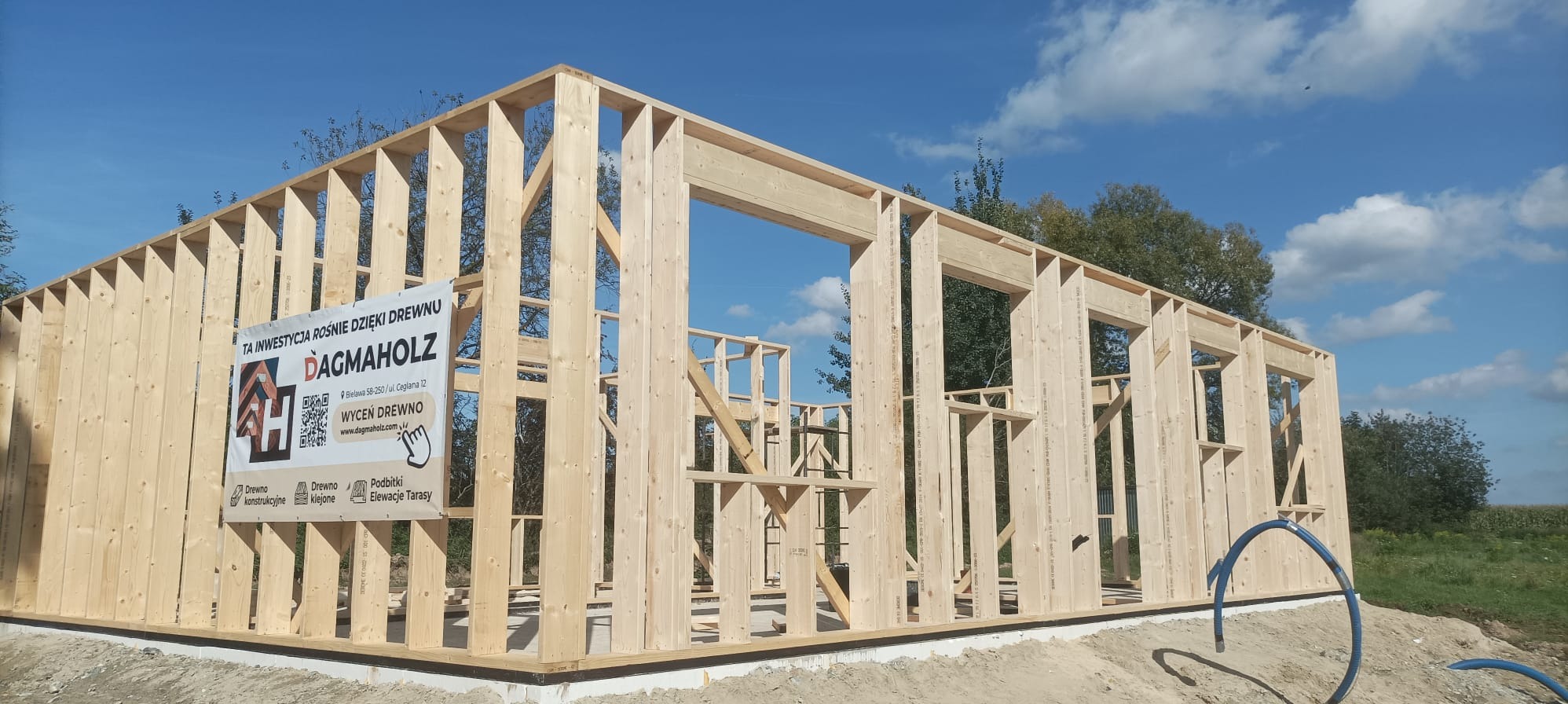 Konstrukcja domu szkieletowego w trakcie budowy, widoczna drewniana rama z logo firmy Dagmaholz na banerze reklamowym. Błękitne niebo w tle.