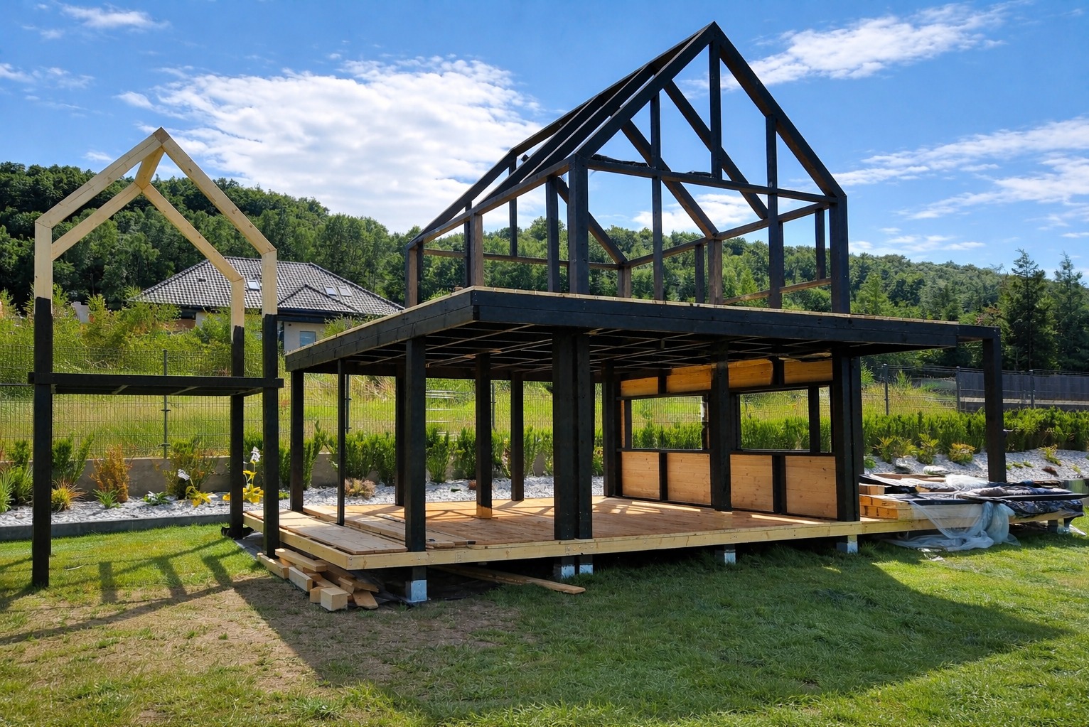 Wznoszona konstrukcja drewniana z czarnymi elementami, prawdopodobnie pergola lub domek letniskowy, na tle zieleni i błękitnego nieba. Widoczne detale konstrukcyjne i etap budowy.
