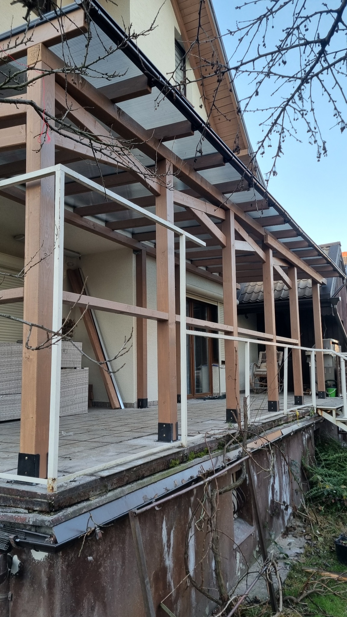 Zadaszony taras z drewnianą konstrukcją i białymi barierkami, widok z dołu. Elementy konstrukcyjne w kolorze brązowym. W tle budynek z oknami i roletami.