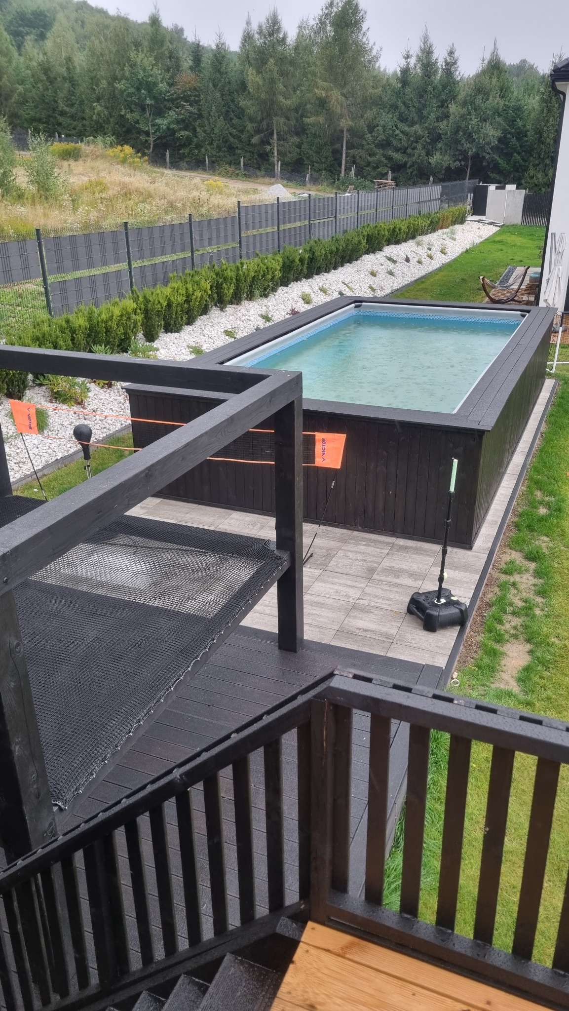 Czarny, drewniany taras z siatką i schodami prowadzący do basenu w ogrodzie. Widok z góry na konstrukcję i otoczenie z zielenią.