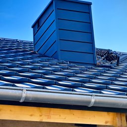 Usługi Ciesielsko Dekarskie - Niebieski komin z blachy na dachu z dachówki ceramicznej, widoczny fragment rynny i ściany budynku w trakcie budowy, błękitne niebo w tle.