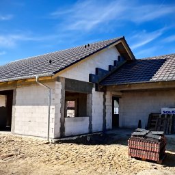 Usługi Ciesielsko Dekarskie - Dom w trakcie budowy z częściowo położonym dachem z dachówki ceramicznej. Widoczne palety z materiałami budowlanymi i niezabudowane otwory okienne.