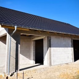 Usługi Ciesielsko Dekarskie - Surowy budynek z czarnym dachem i rynnami w słoneczny dzień. Widoczne bloczki betonowe i drewniana konstrukcja dachu.