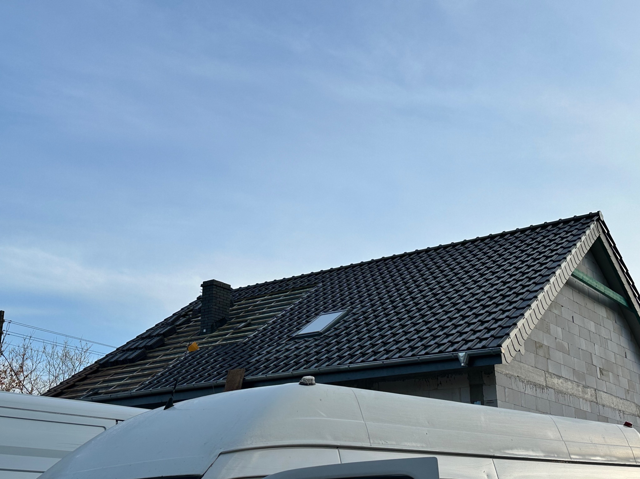 Częściowo pokryty dach z czarną dachówką, widoczne elementy więźby i okno dachowe. W tle błękitne niebo i fragment białego busa.