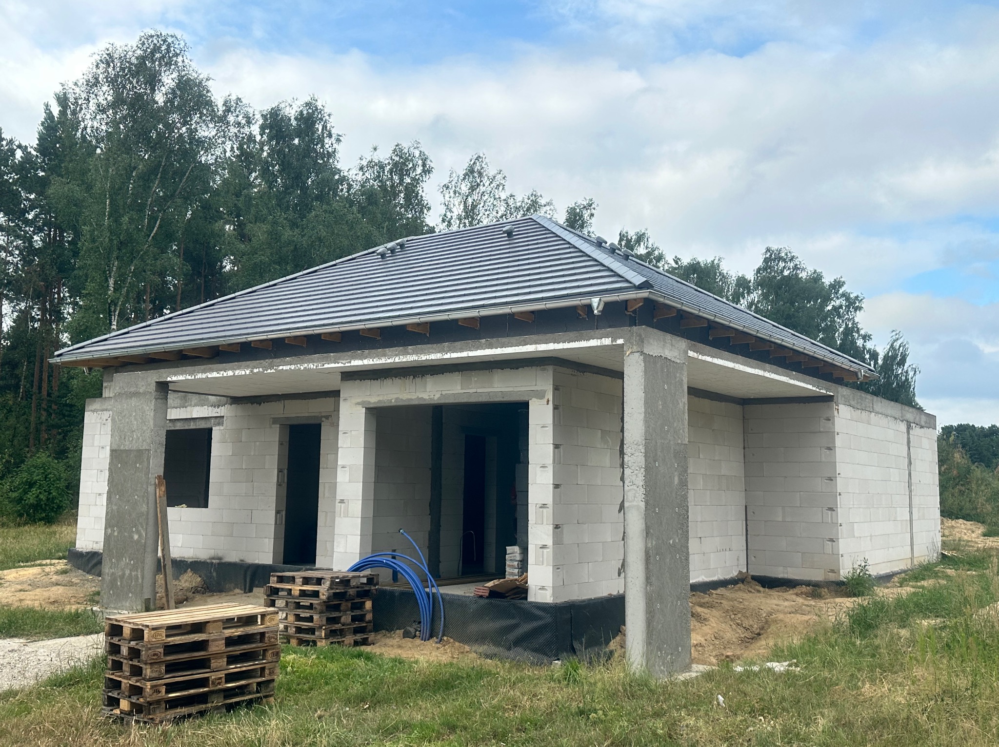 Surowy stan domu jednorodzinnego z szarym dachem z dachówki ceramicznej, widoczne palety i niebieskie rury na tle zieleni. Budowa w toku, stan surowy otwarty.