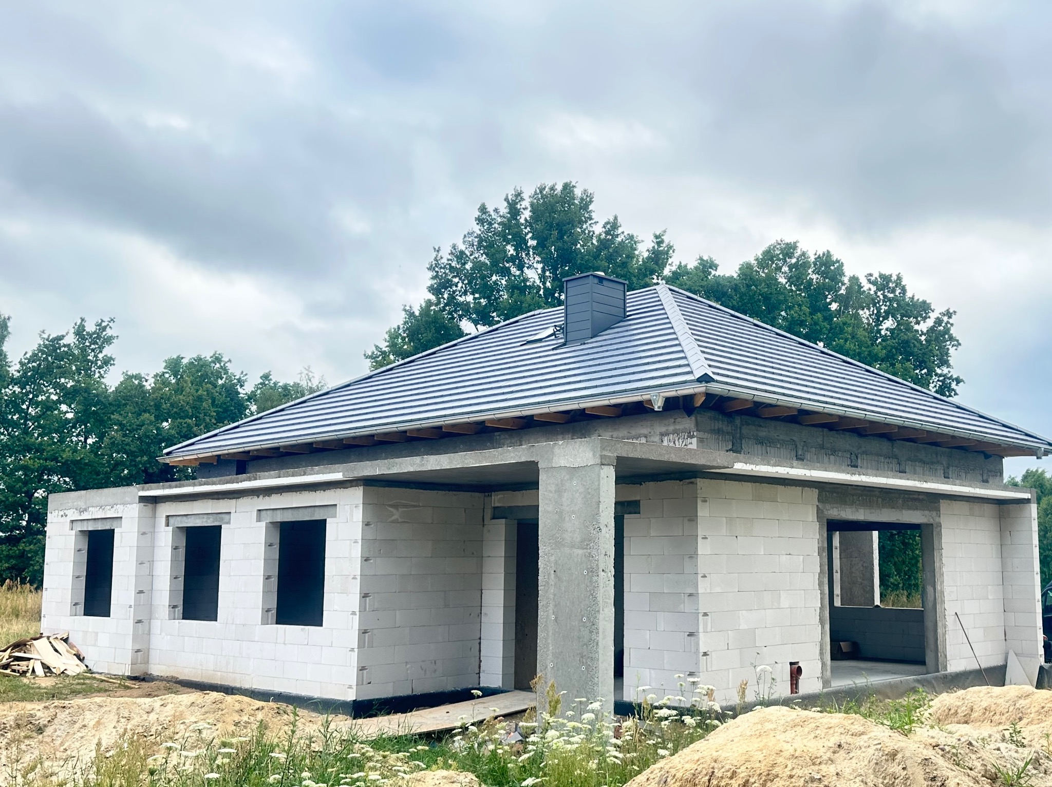Dom w trakcie budowy z szarym dachem z dachówki ceramicznej i kominem. Ściany z białych bloczków, widoczne otwory okienne i drzwiowe. Tło: drzewa i pochmurne niebo.