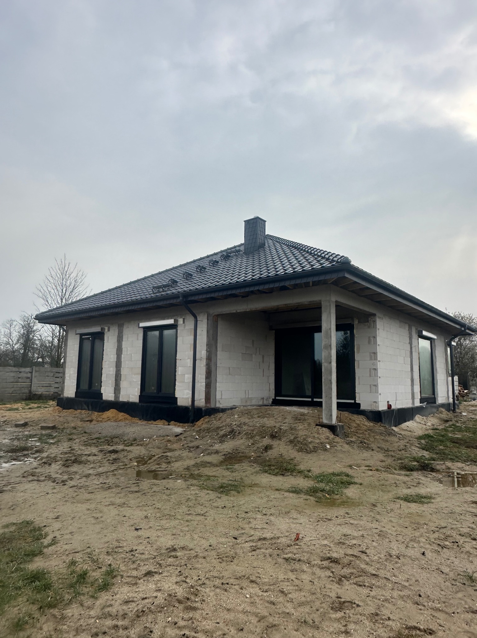Dom jednorodzinny w trakcie budowy, z widocznym dachem pokrytym ciemną dachówką i surowymi ścianami z betonu komórkowego. Widoczne czarne ramy okienne i przygotowany teren.
