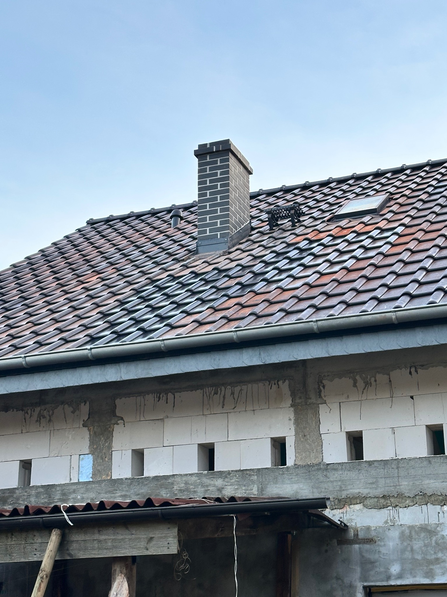 Dach z dachówką karpiówką w odcieniach brązu i czerwieni, z nowym, ceglanym kominem i oknem dachowym. Widoczne obróbki blacharskie i fragment ściany budynku w trakcie budowy.